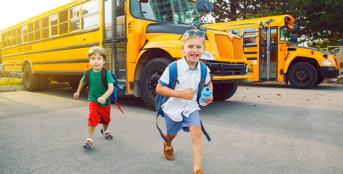 Two young boys running playfully in front of a yellow school bus on a sunny day.
