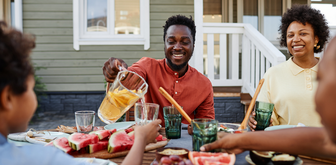 A family smiles while enjoying food outdoors, with refreshing beverages and watermelon.